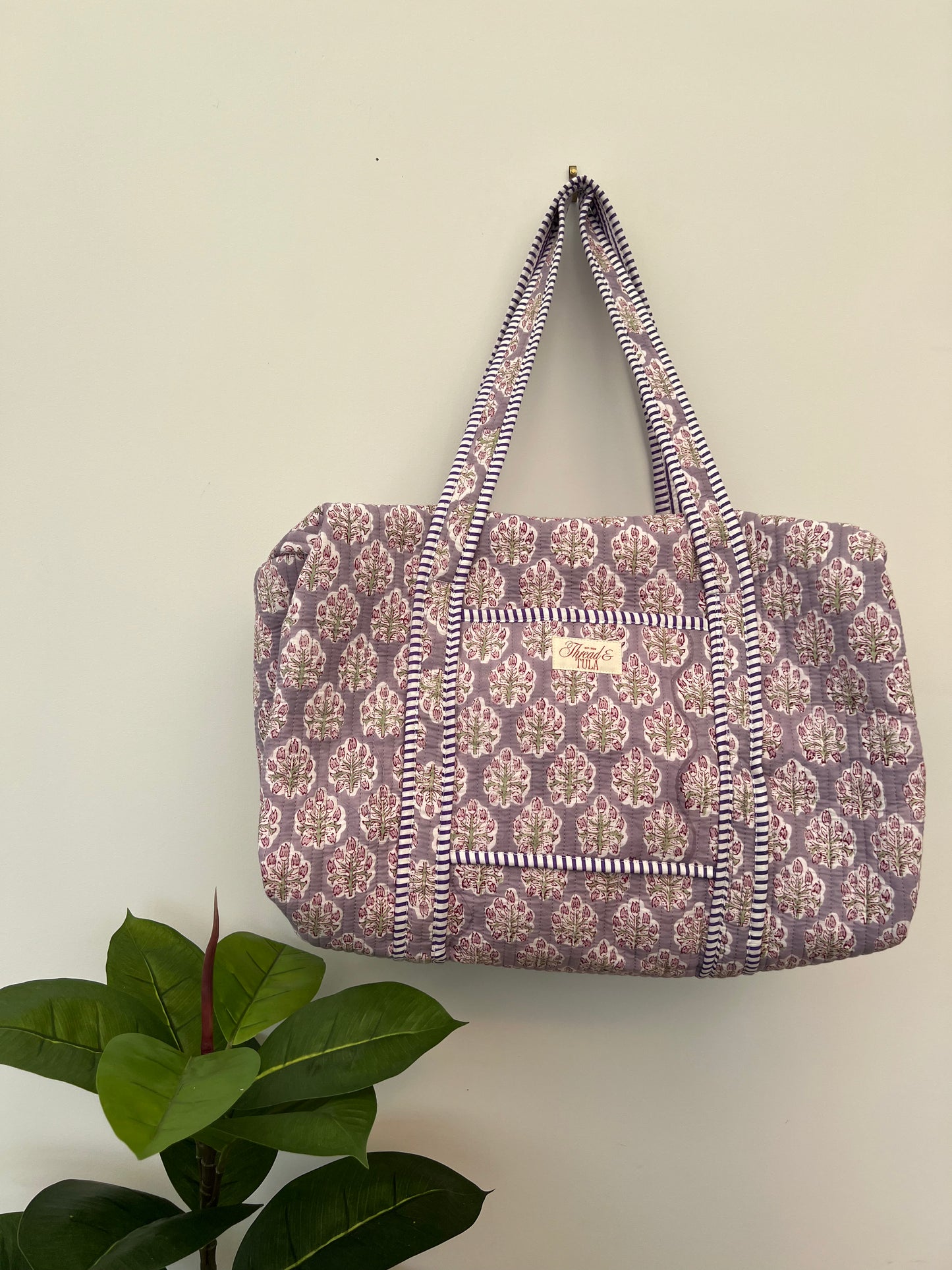 A fabric tote bag with a purple and beige floral block print pattern and striped handles, hanging on a plain beige wall with a green plant leaf in the lower left corner.