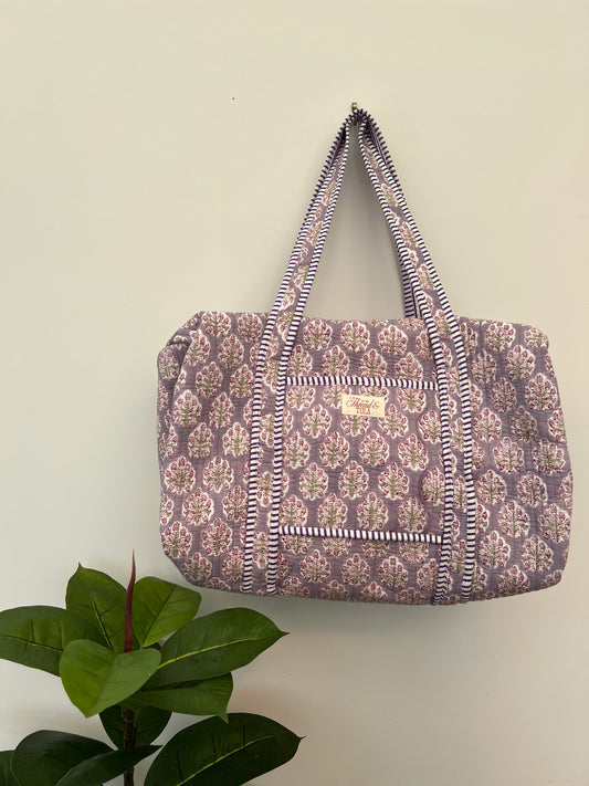 A fabric tote bag with a purple and beige floral block print pattern and striped handles, hanging on a plain beige wall with a green plant leaf in the lower left corner.